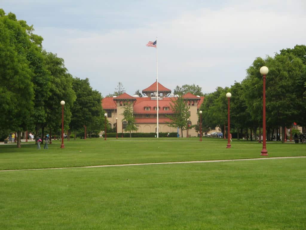 A broad green lawn with paths and lampposts leading to a red-roofed, Spanish-style college building with a flagpole, framed by leafy trees.