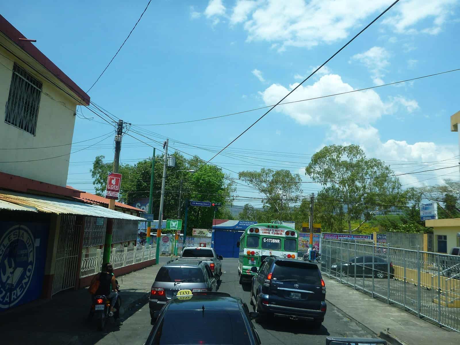 A busy street in Escuintla, Guatemala, with cars, a green bus and overhead power lines under a blue sky