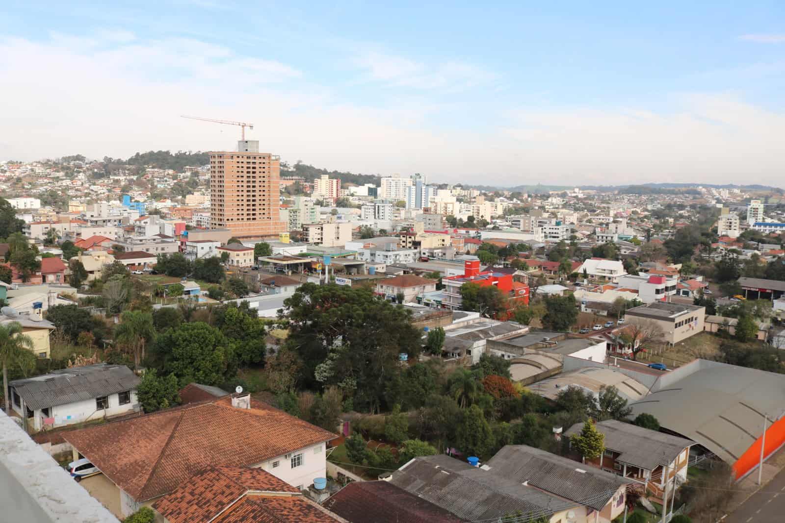 An elevated view over the rooftops, trees and apartment buildings of a small hillside city under a pale blue sky