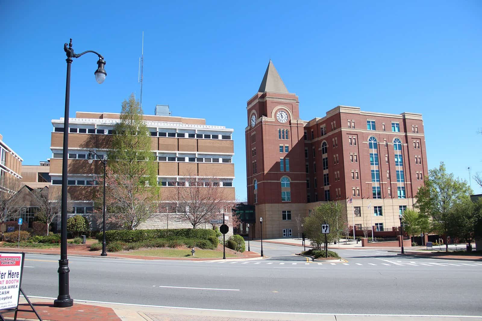 A red brick courthouse with a clock tower beside an older office building, across an empty street under a clear blue sky