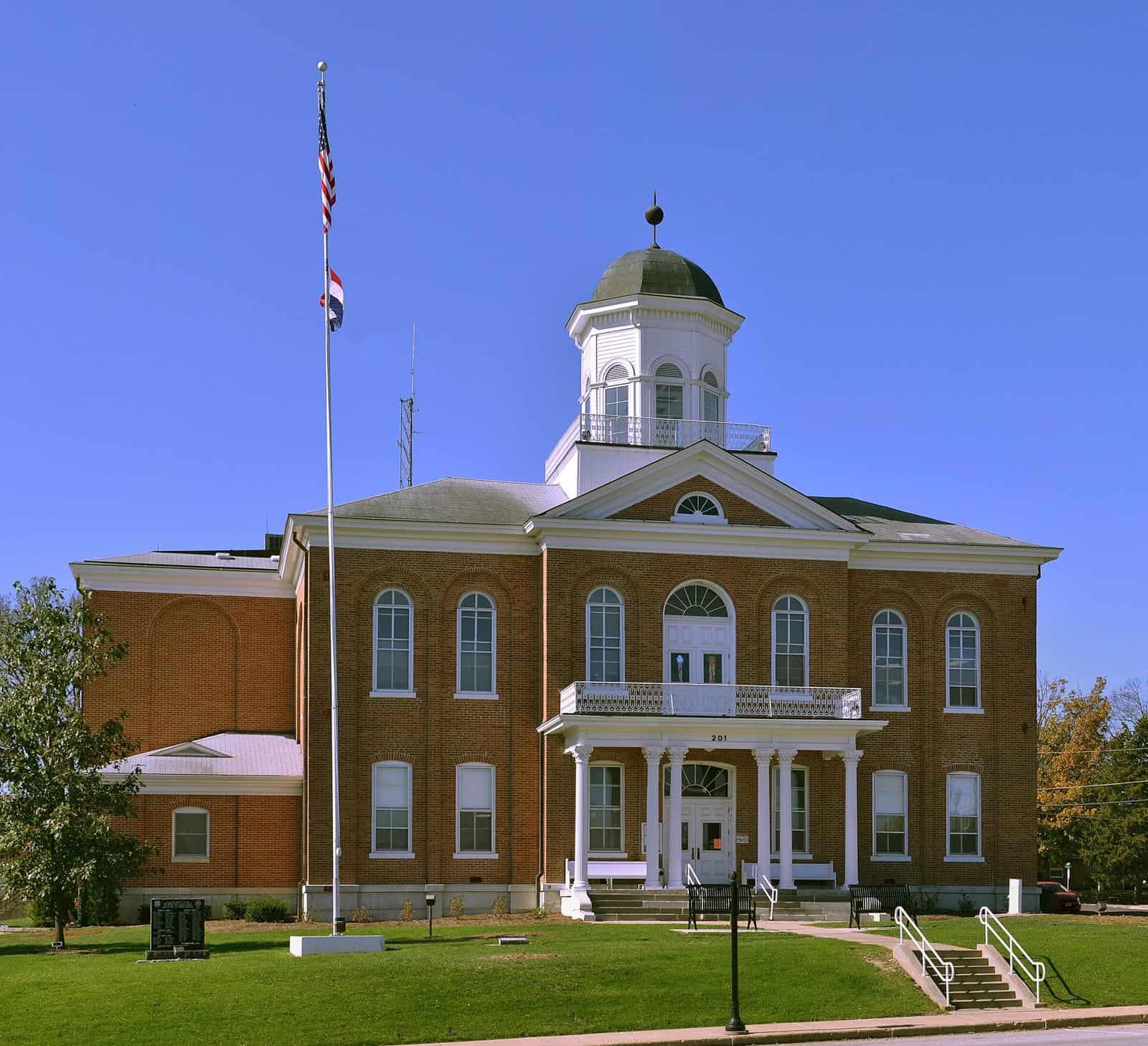 A two-story red brick courthouse with white columns, a balcony and a domed cupola, with a flagpole on a green lawn under a clear blue sky