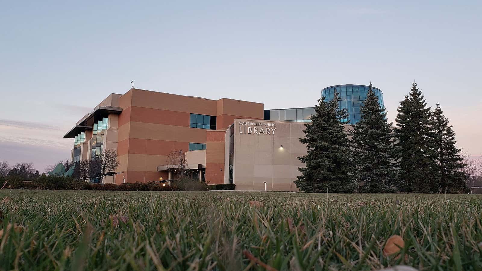 A modern brick and stone library building with a curved glass tower, evergreen trees and a lawn in the foreground at dusk