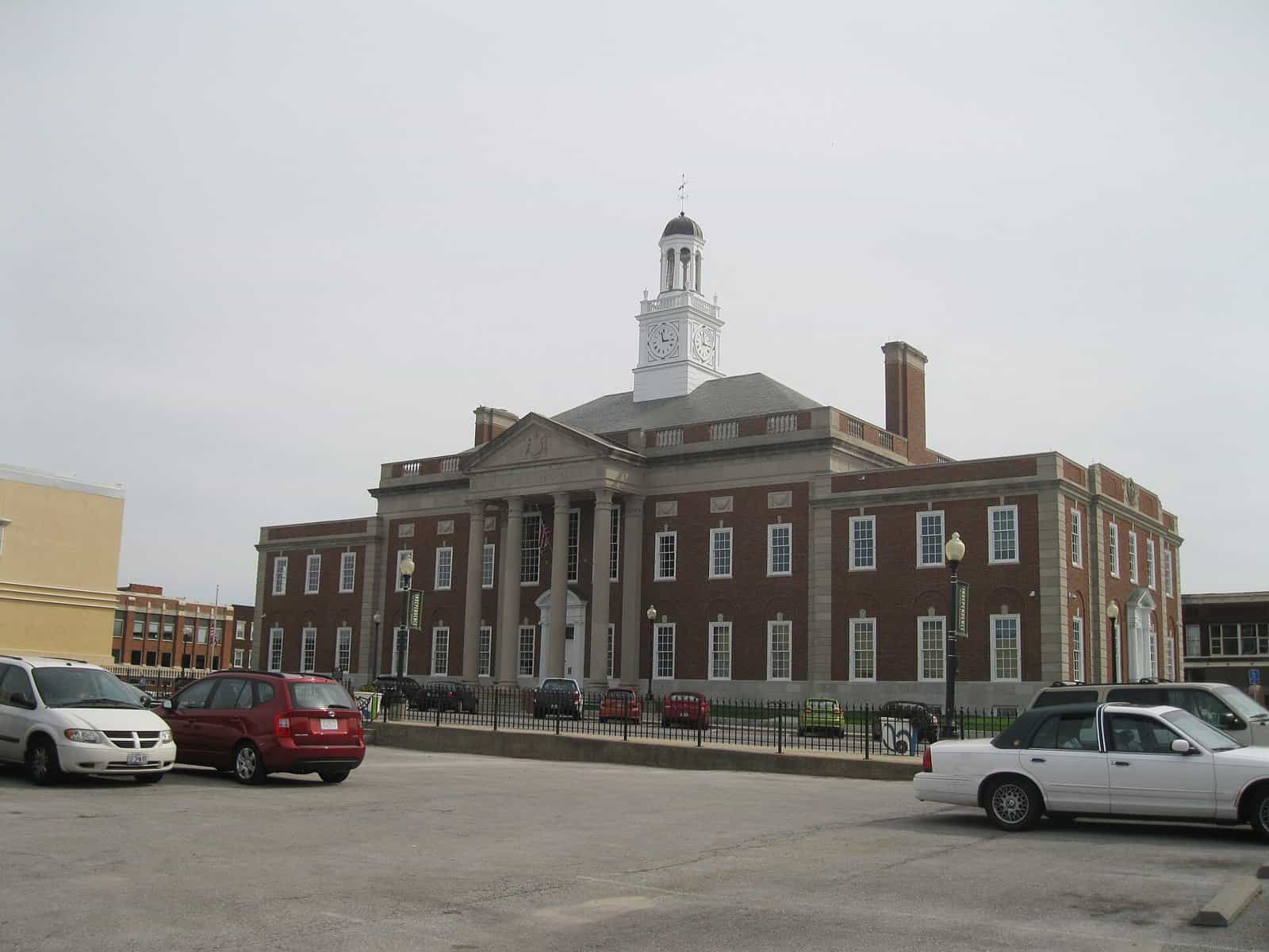 A red brick courthouse with white columns and a white clock cupola, seen across a parking lot with parked cars under a gray sky