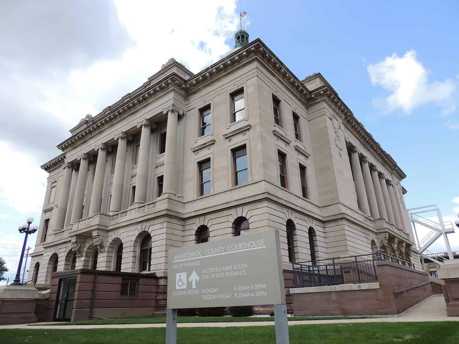A pale stone Neoclassical courthouse with tall columns and a small green cupola, with a sign reading Manitowoc County Courthouse in front