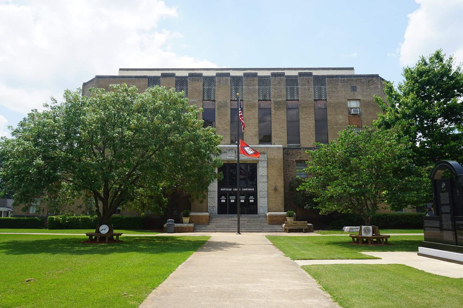 A tan brick Art Deco courthouse with tall vertical windows, an American flag and an Arkansas flag on a pole, and a lawn and walkway in front