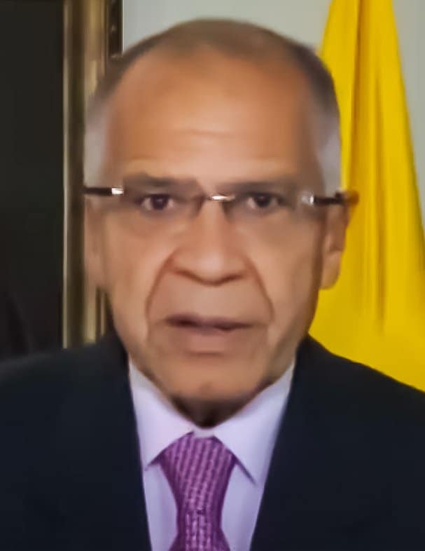 Alfonso Gómez Méndez, a grey-haired man with glasses in a dark suit and purple tie, speaking in front of a Colombian flag.