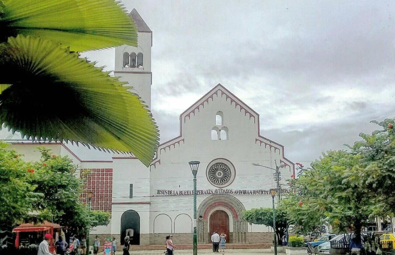 A white church with a tall bell tower in Chone, Ecuador, framed by palm leaves under a cloudy sky.
