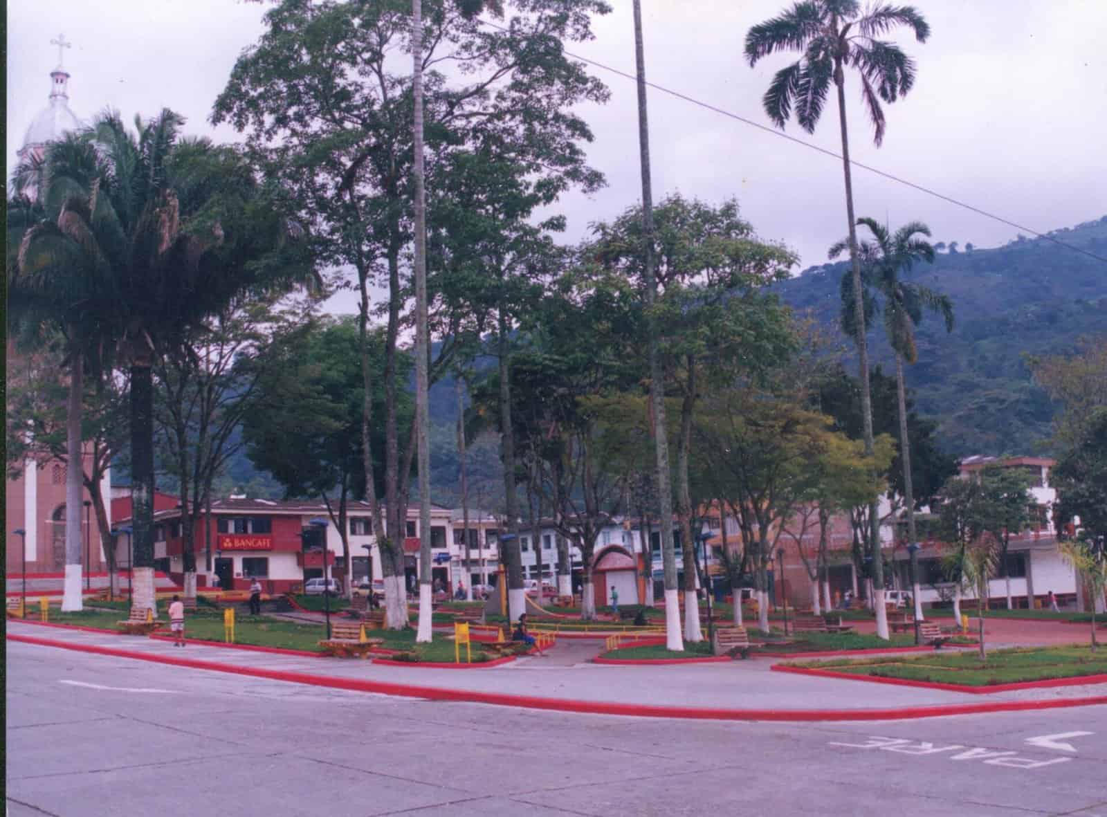 The main square of Génova, a small Andean town, with palm trees, a church spire and green mountains behind.
