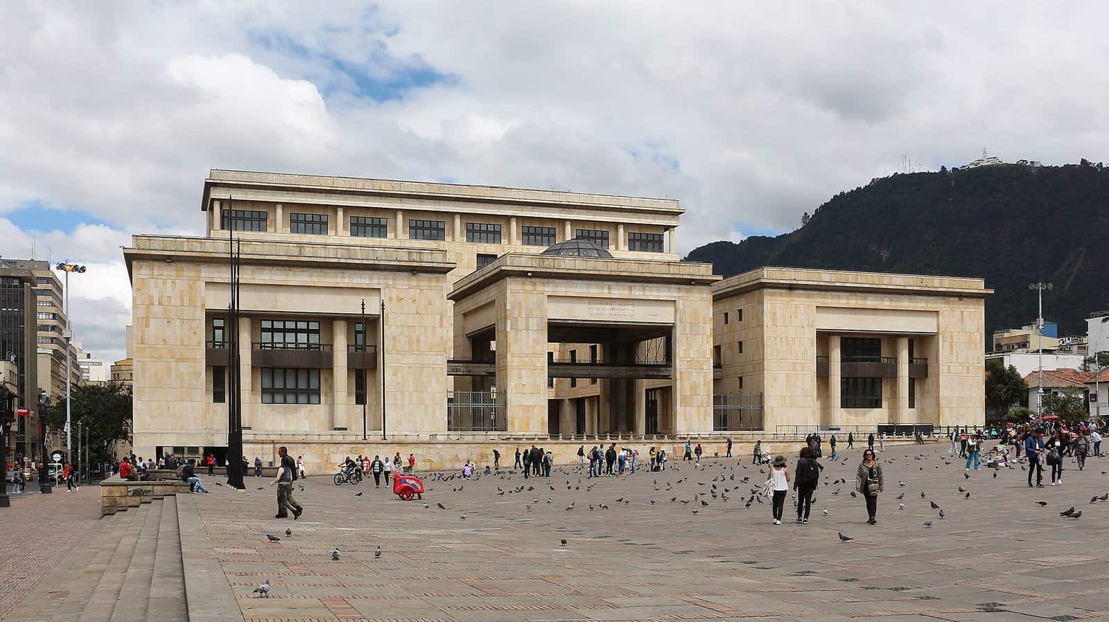 The Palacio de Justicia in Bogotá, a long pale stone building with square columns, across a wide plaza with pigeons, below green mountains.