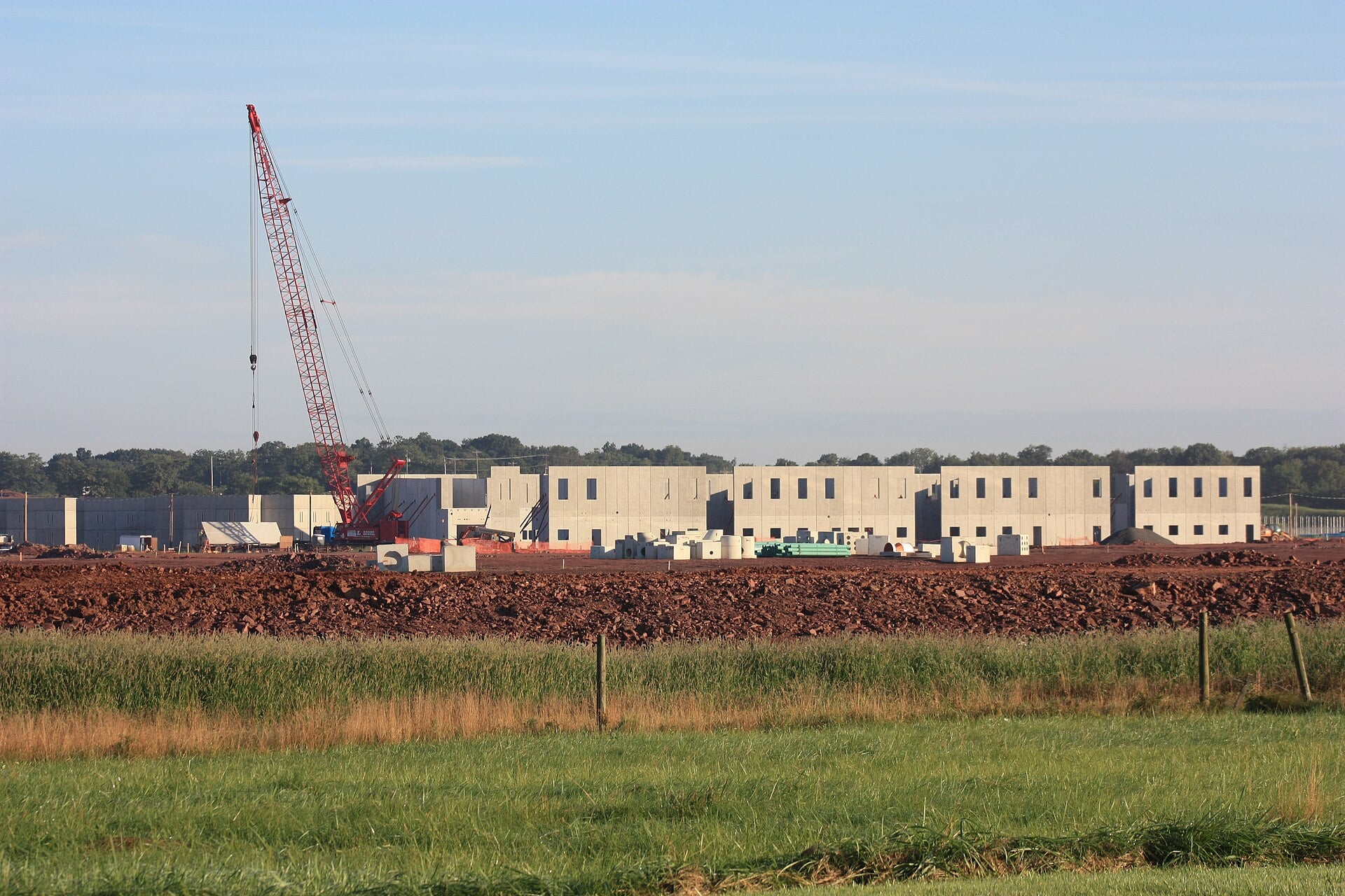 Concrete prison buildings under construction behind a field, with a tall red crane at left