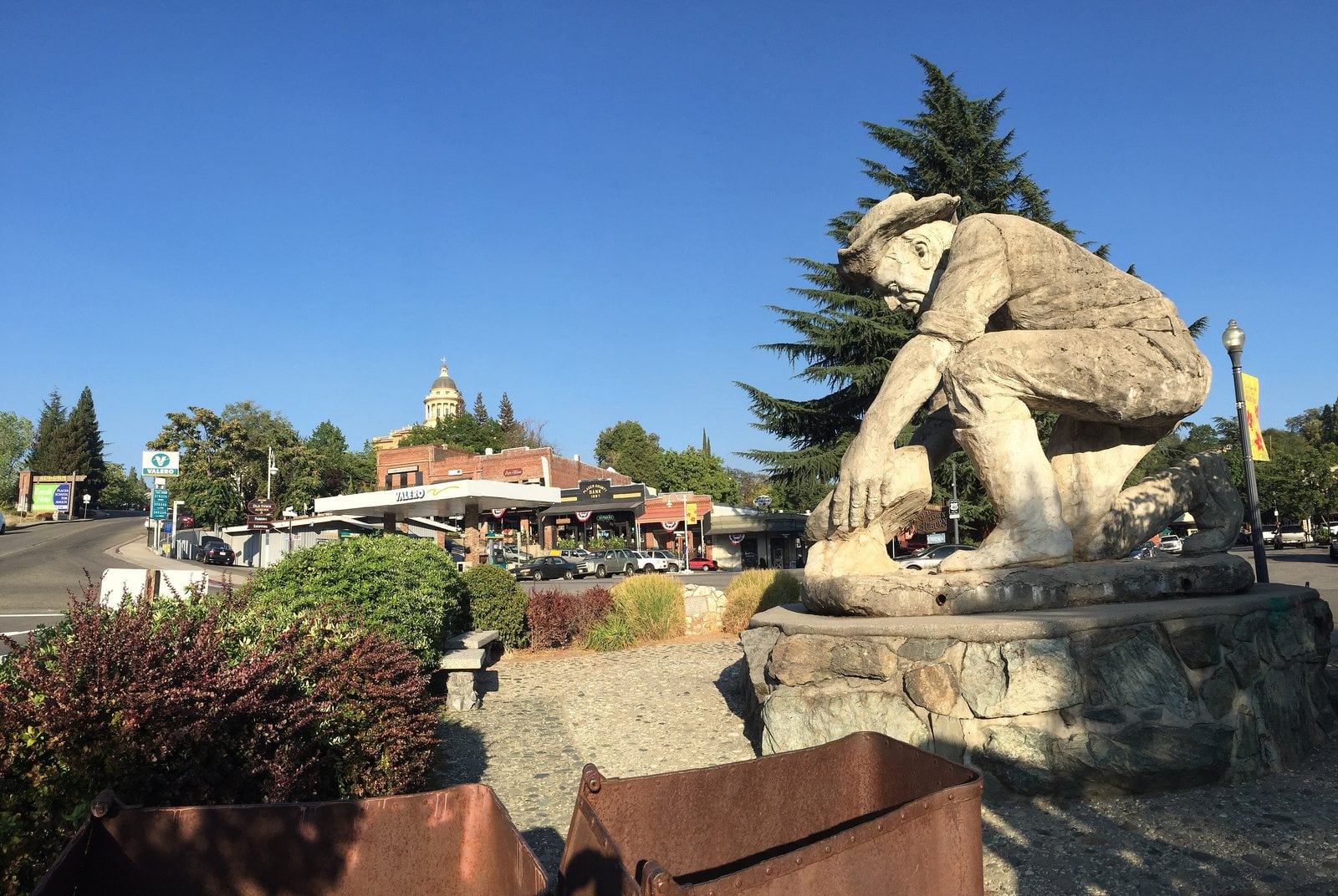 Downtown Auburn, California, with a large sculpted stone figure on a hillside garden in the foreground and historic buildings and trees behind.