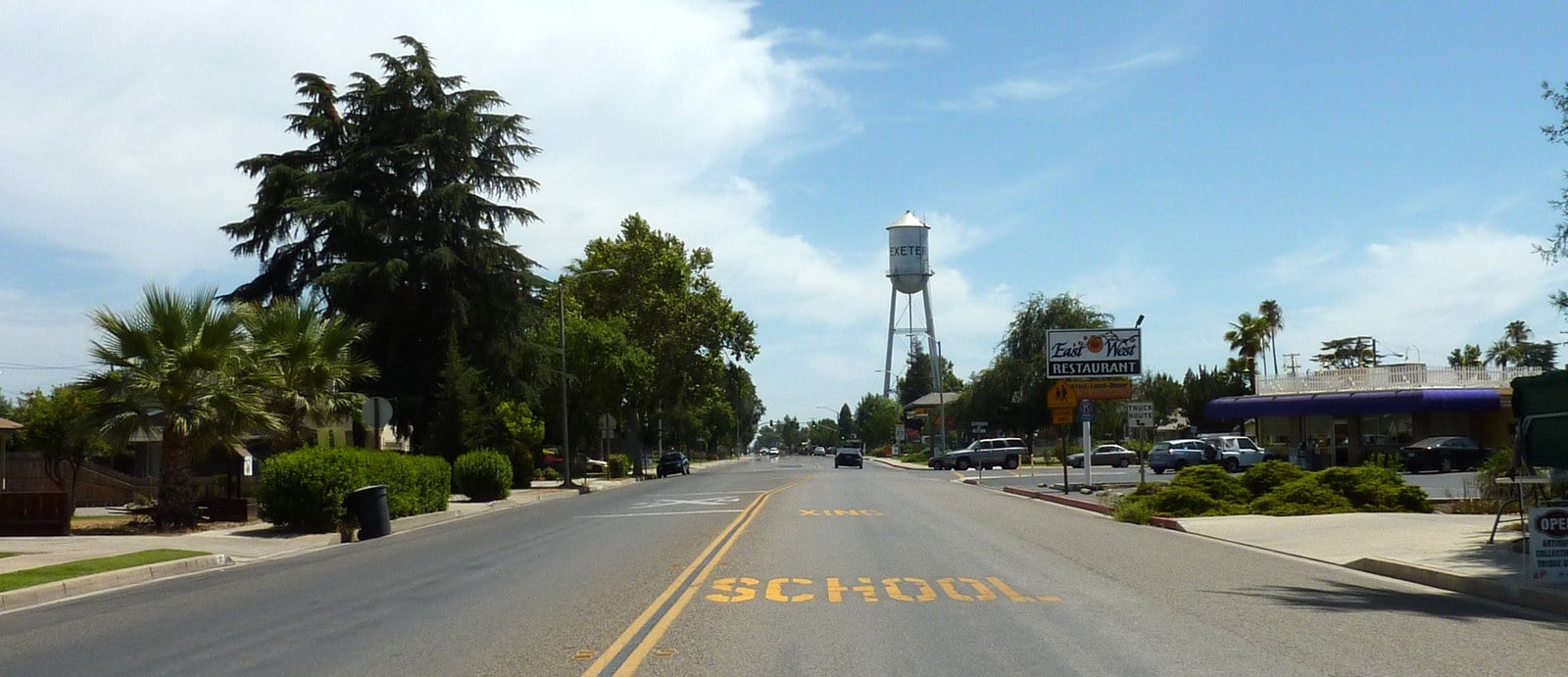 A quiet two-lane road through Exeter, California, lined with trees and low buildings, with a water tower visible in the distance.