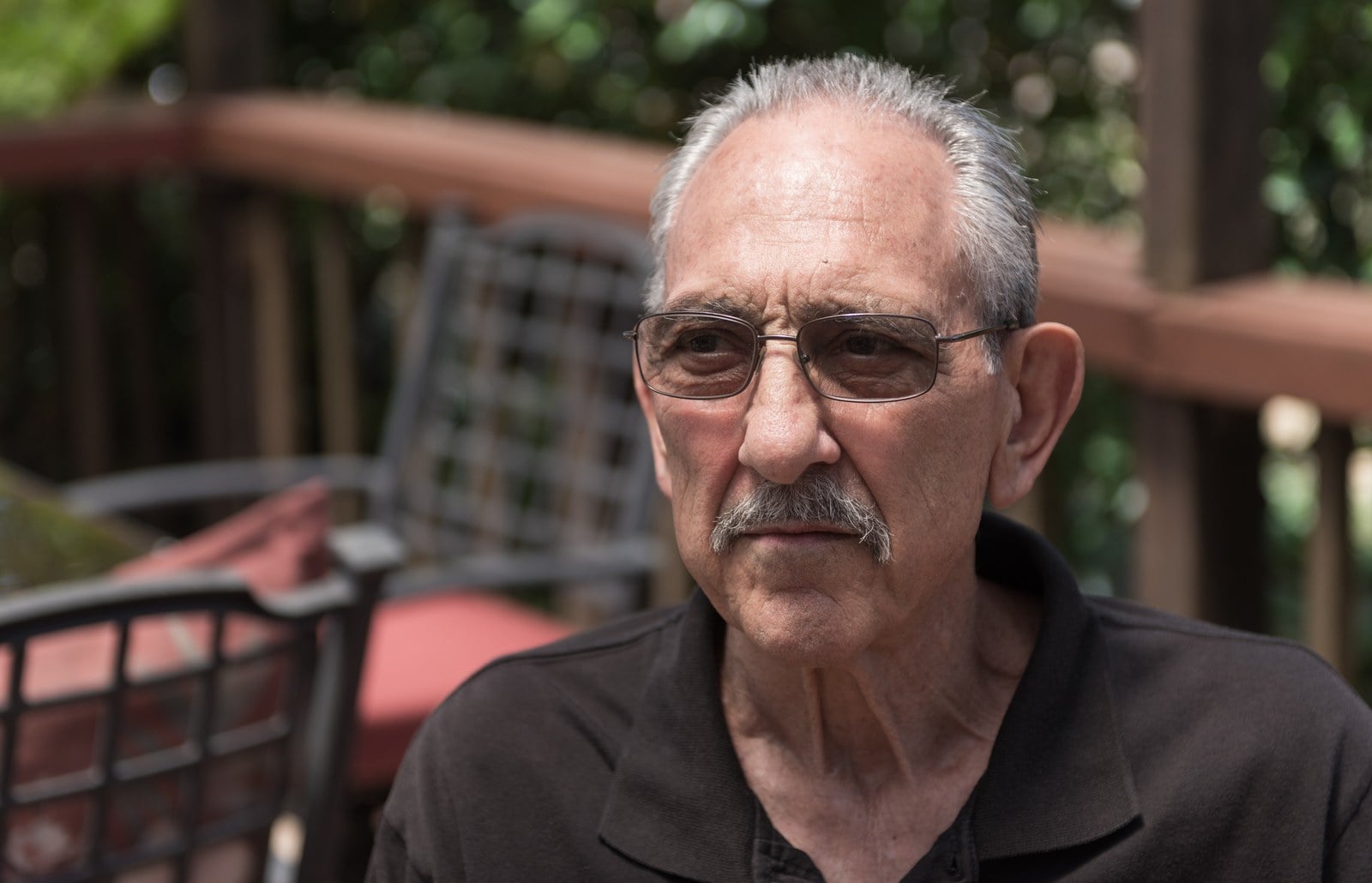 An older man with white hair and glasses, wearing a dark shirt, seated outdoors on a porch with trees behind him.