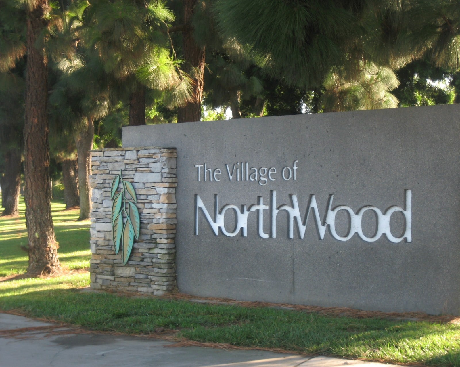 A low stone wall bearing the words The Village of Northwood in raised gray letters, framed by shrubs and tall eucalyptus trees in Irvine.