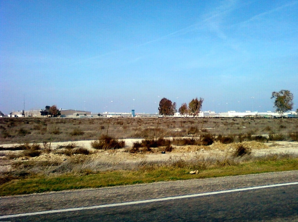 A distant view across flat dry ground of the low buildings and perimeter fencing of North Kern State Prison in Delano, California, under a pale blue sky.