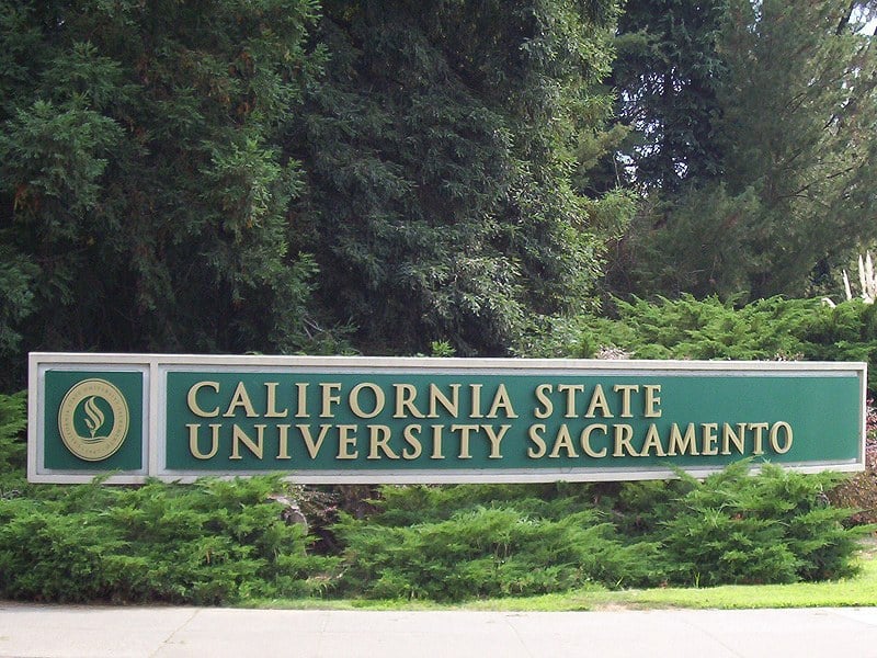 A green sign reading California State University Sacramento set in a low brick wall at a campus entrance, with trees behind it.