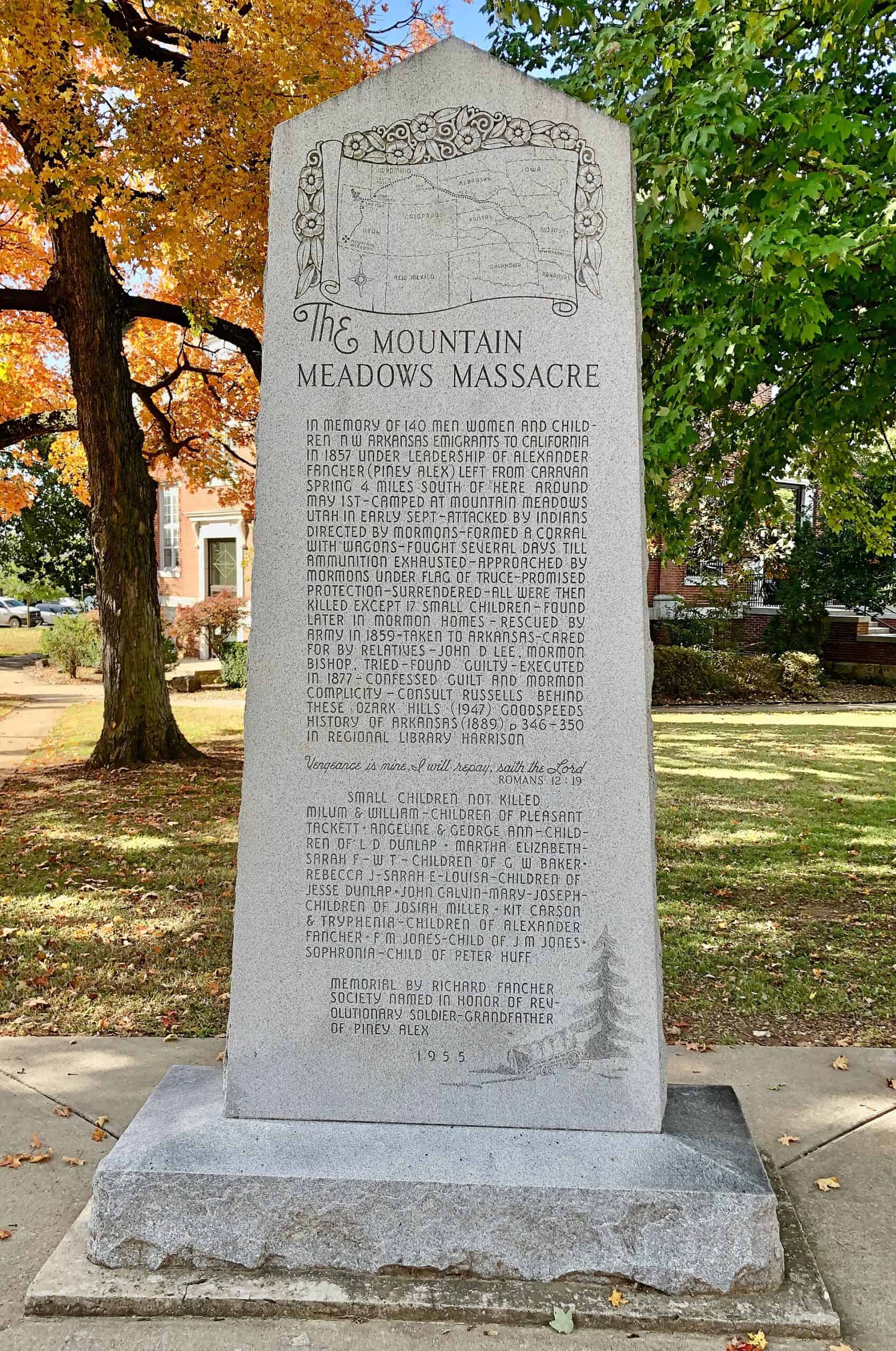 A tall granite monument with an engraved map and inscription standing on a lawn in a town square, with autumn trees behind it