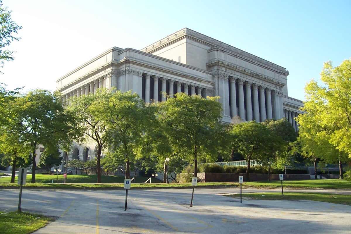 The Milwaukee County Courthouse, a long neoclassical stone building with tall columns, seen across a parking lot and trees