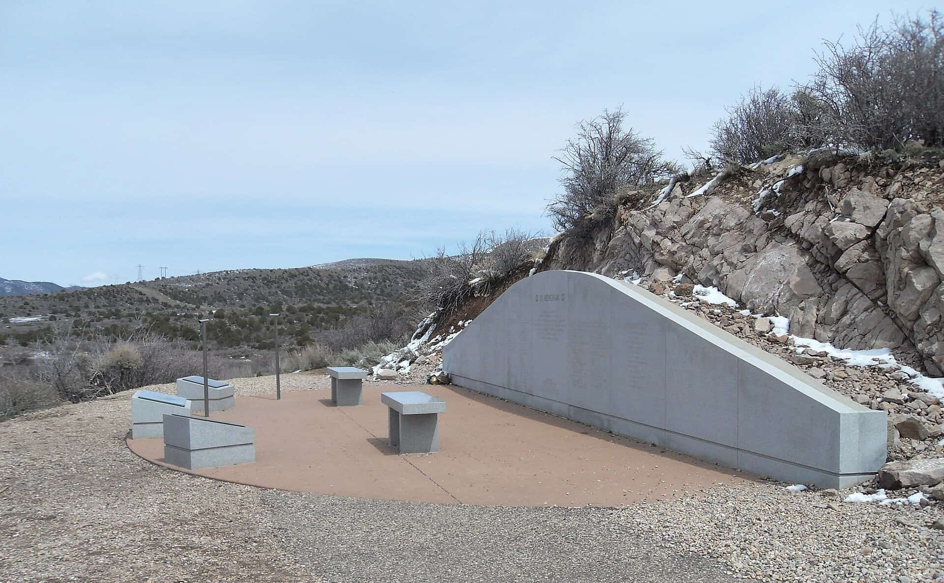 A curved grey stone memorial wall set into a rocky hillside above a paved circle with stone benches