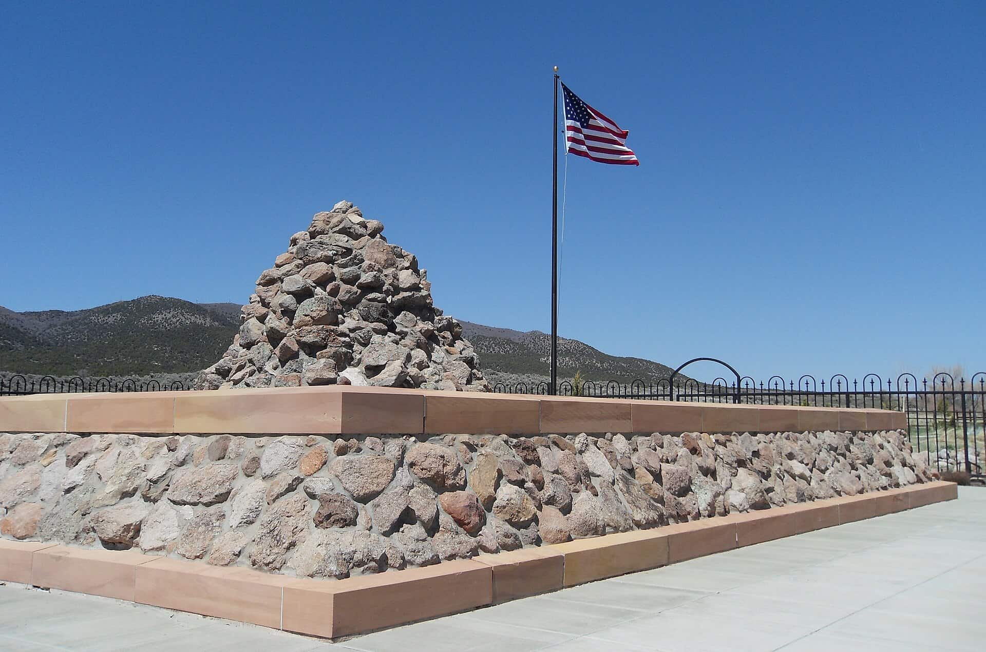 A conical stone cairn rising from a low rectangular stone wall, with an American flag on a pole and hills under a clear sky