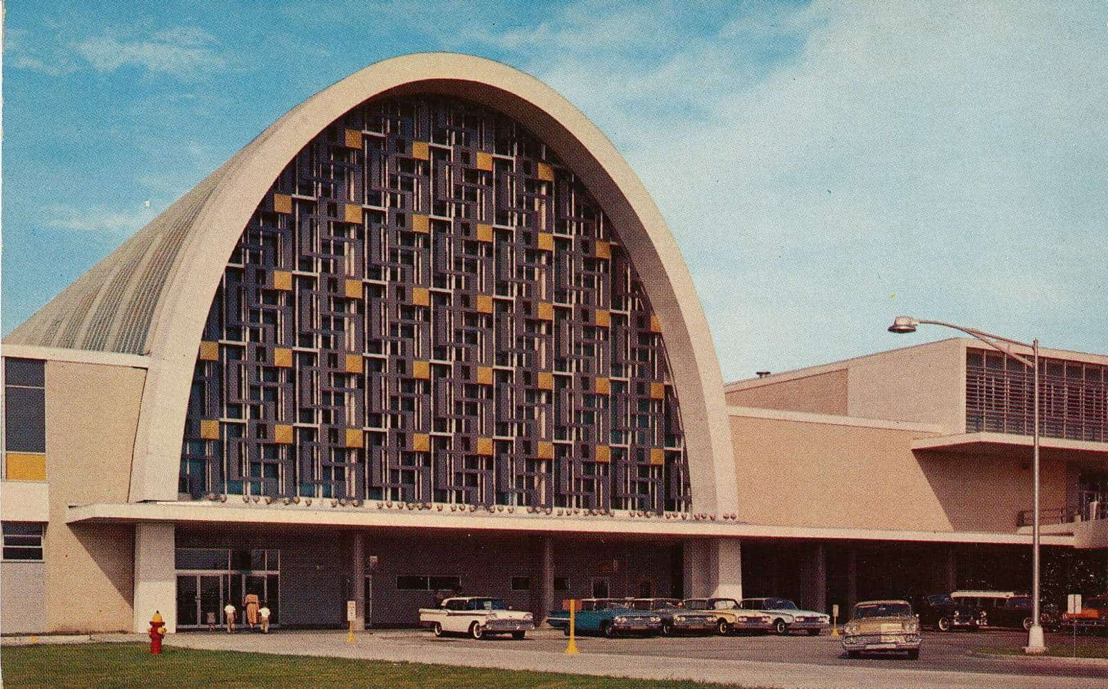 Vintage color postcard of an airport terminal with a tall arched facade of patterned glass, with 1950s and 1960s cars parked along the entrance