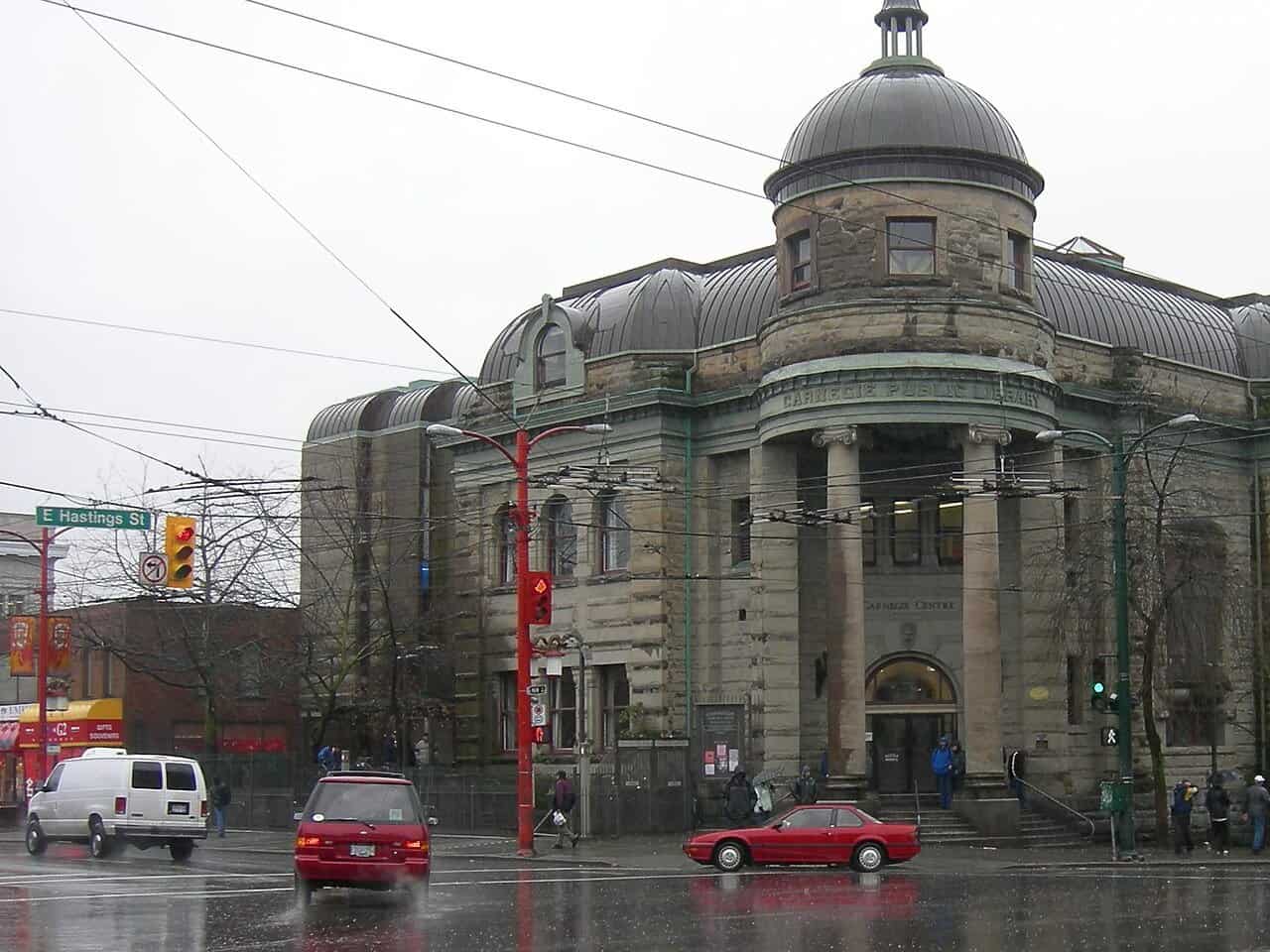 The Carnegie Centre, a domed sandstone former library with columns, on a rainy corner of East Hastings Street in Vancouver, with cars passing.