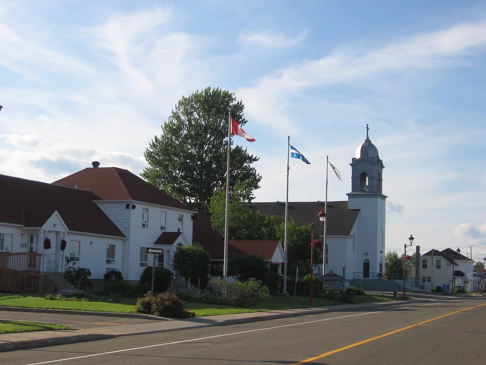 A white clapboard city hall and a white church with a domed bell tower, behind flagpoles flying the Canadian and Quebec flags, beside a quiet road.
