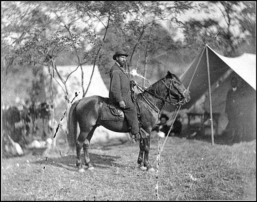 A bearded man in a dark coat and hat sits on a horse in an open field, with tents and trees behind him