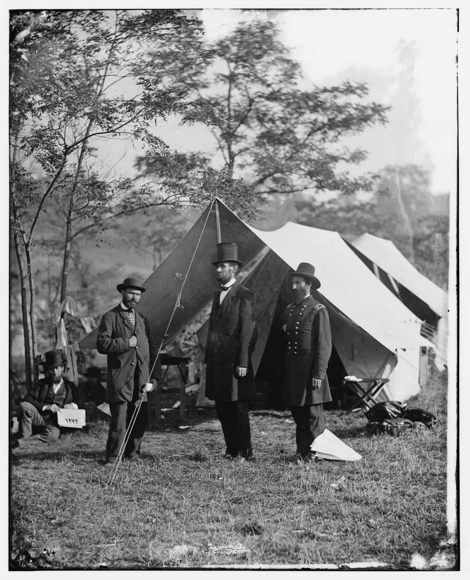 Three men stand in front of a canvas tent in a grassy army camp: a bearded man in a dark coat and low hat on the left, a very tall man in a black suit and stovepipe hat in the centre, and a uniformed officer on the right