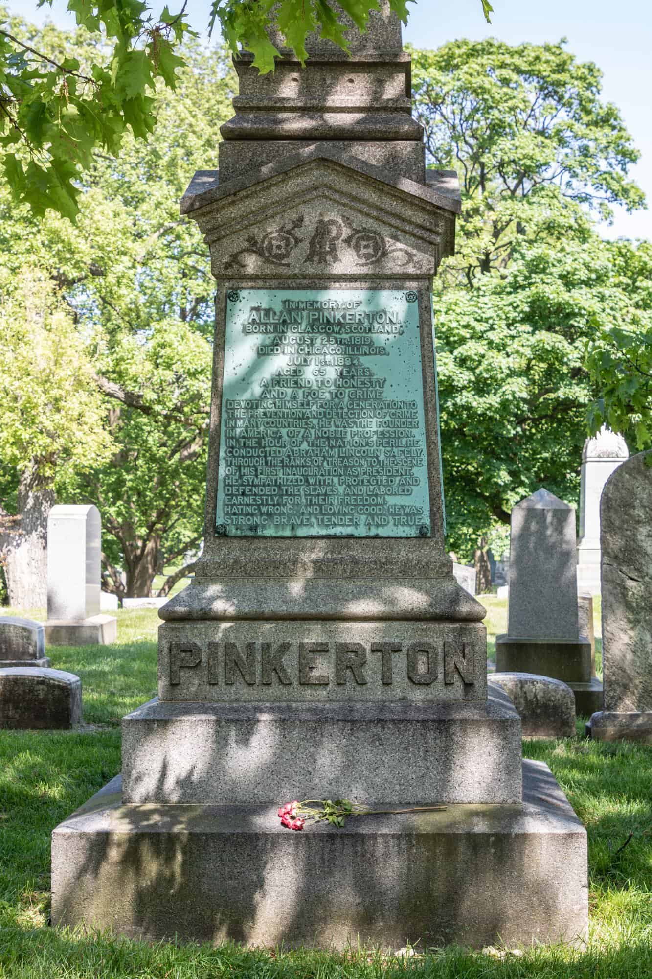 A tall granite monument in a cemetery with a bronze plaque near its top and the name PINKERTON carved into the base, surrounded by grass and trees